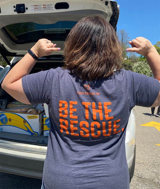 A Food Rescue US volunteer displays the message Be the Rescue on the back of her t-shirt with a car full of food in the background