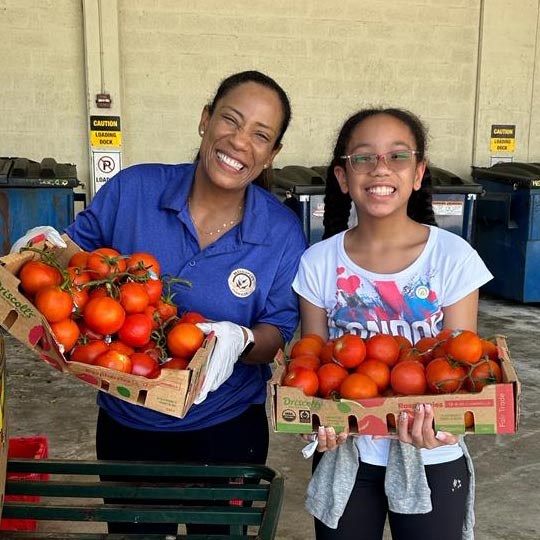 A woman and teen girl smile and hold boxes of fresh tomatoes