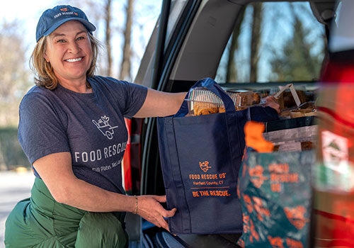 A volunteer smiles at the camera as she unloads bags of food from her car